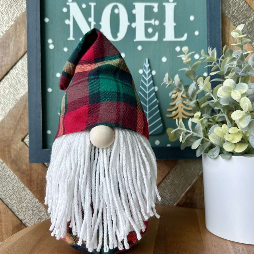 Gnome made without sewing, wearing a red plaid hat standing front of a sign that says NOEL with a plant on the side