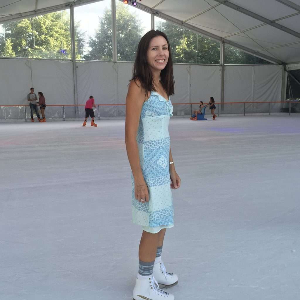 Nicole bolin in a blue dress and ice skates, standing on an ice skating rink