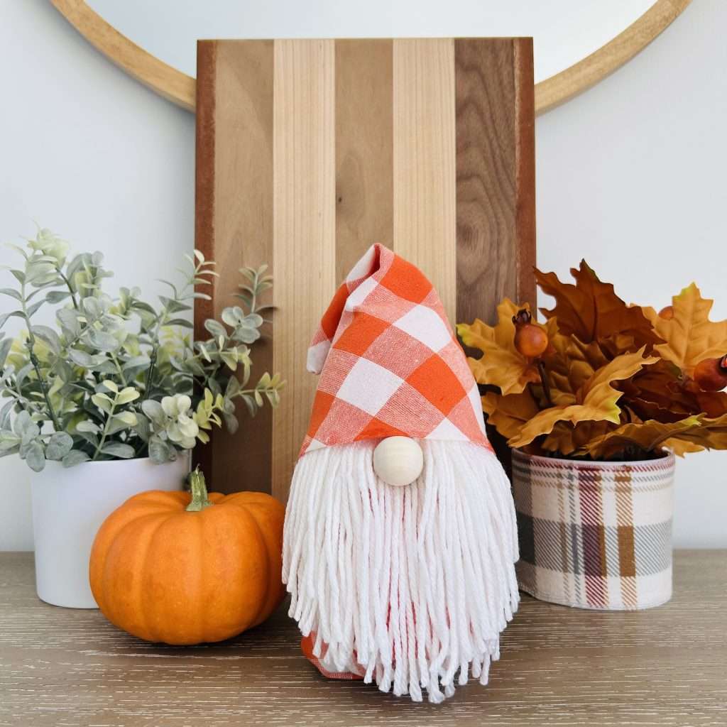 Gnome made without sewing, wearing an orange checkered gingham hat, on a shelf with a pumpkin and plants