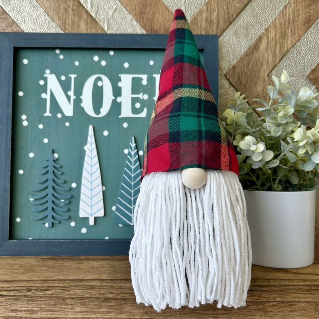 Gnome made without sewing, wearing a pointy red plaid hat standing front of a sign that says NOEL
