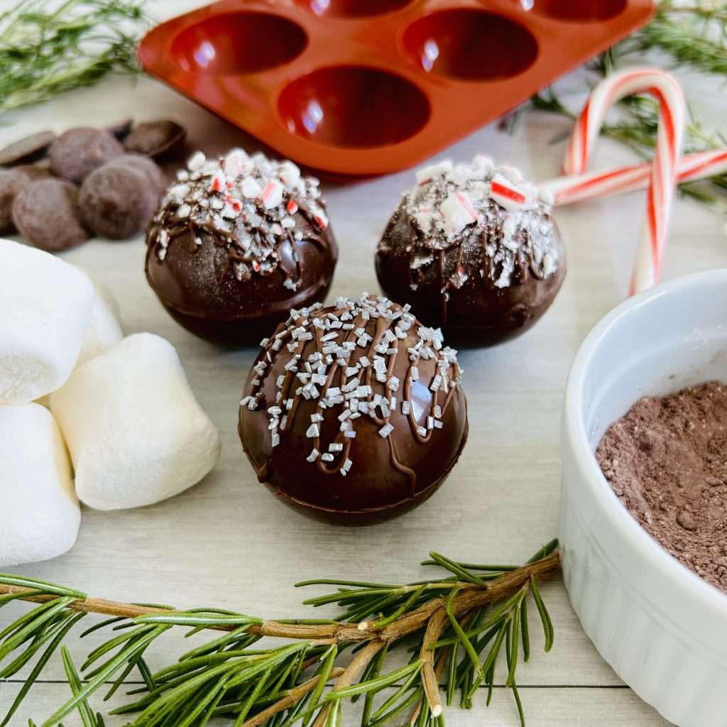3 hot cocoa bombs with silver sprinkles and candy cane sprinkles surrounded by chocolate wafers, marshmallows, hot cocoa mix, candy canes, rosemary branches and a silicon candy mold.
